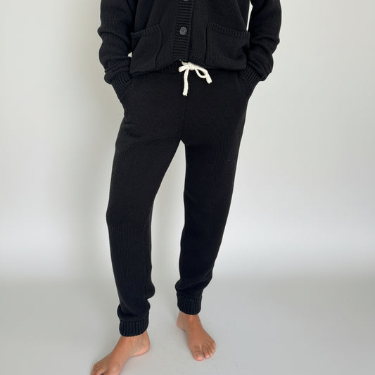 Black knitted cardigan and knitted jogger pants worn by a person on a white background