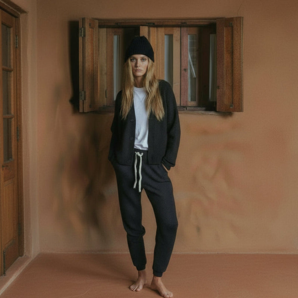 Person wearing a black beanie, black knitted cardigan, white t-shirt, and black knitted pants standing in front of a wooden wall.