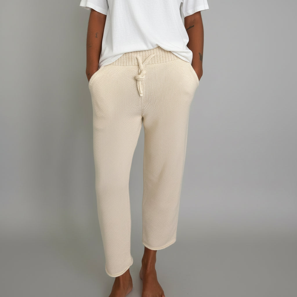 Woman wearing a white t-shirt and ecru pants in a room with a grey/white backdrop and tiled floor.