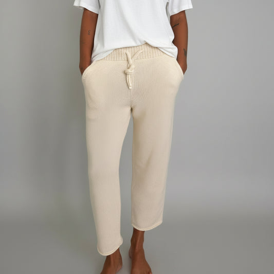 Woman wearing a white t-shirt and ecru pants in a room with a grey/white backdrop and tiled floor.